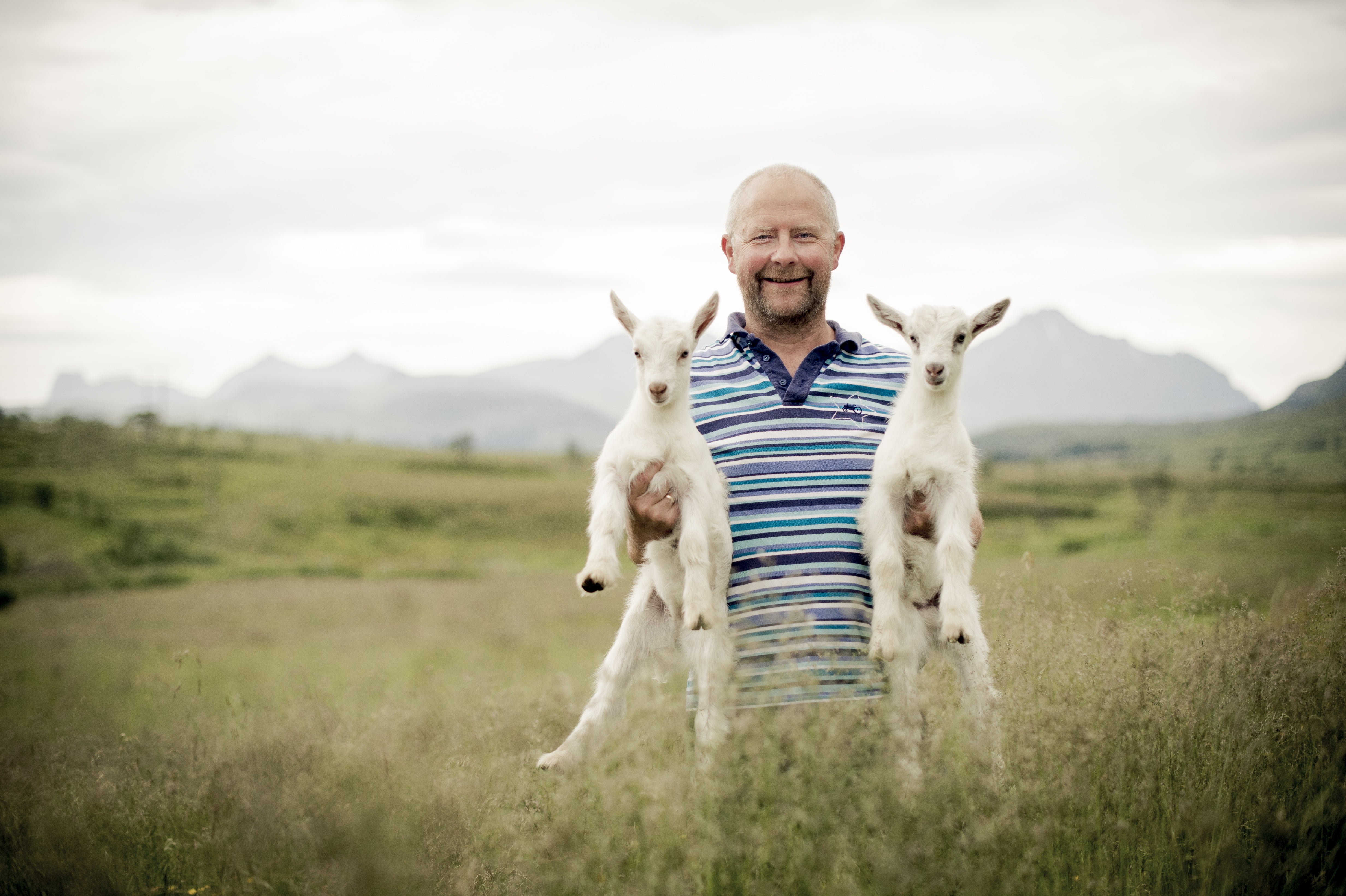 Sauebonde står på en gresslette og holder et lam under hver arm.